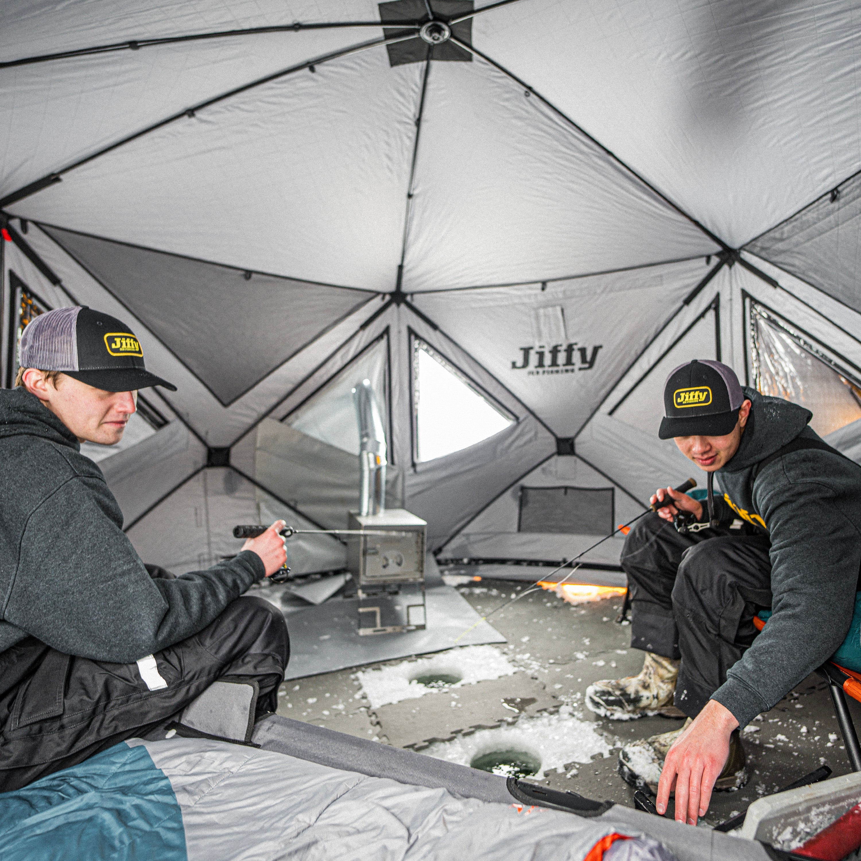 Two people inside a Jiffy tent, both sitting holding ice fishing rods, with a stove and cots visible.